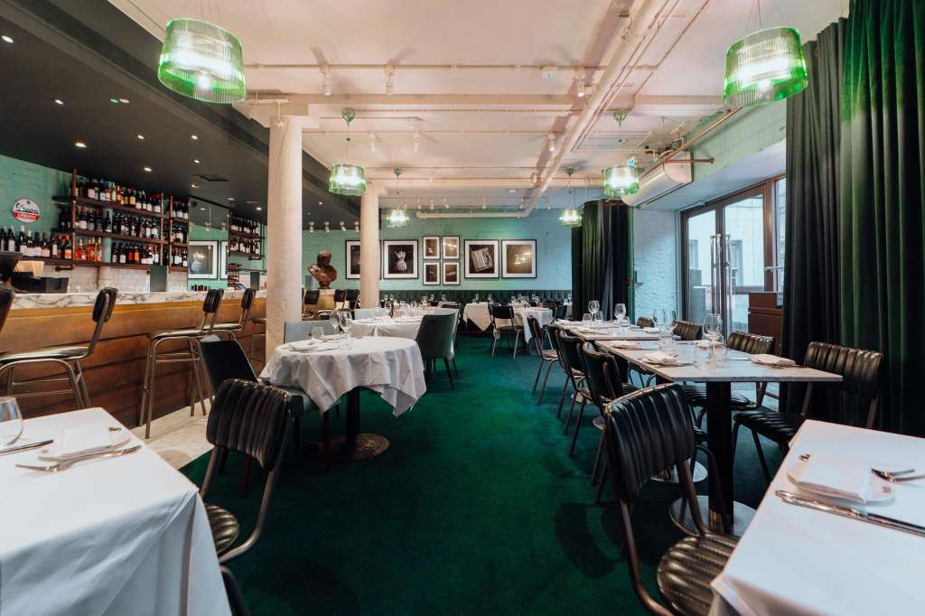 Dining room with round and rectangular tables set with white linens, black chairs, green carpet, and bar area.