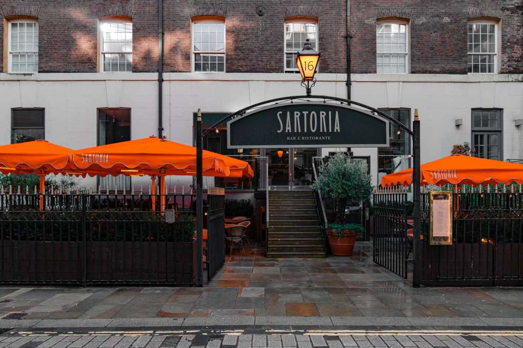 Sartoria bar and restaurant exterior with orange umbrellas, black railings, and brick facade on a London street.