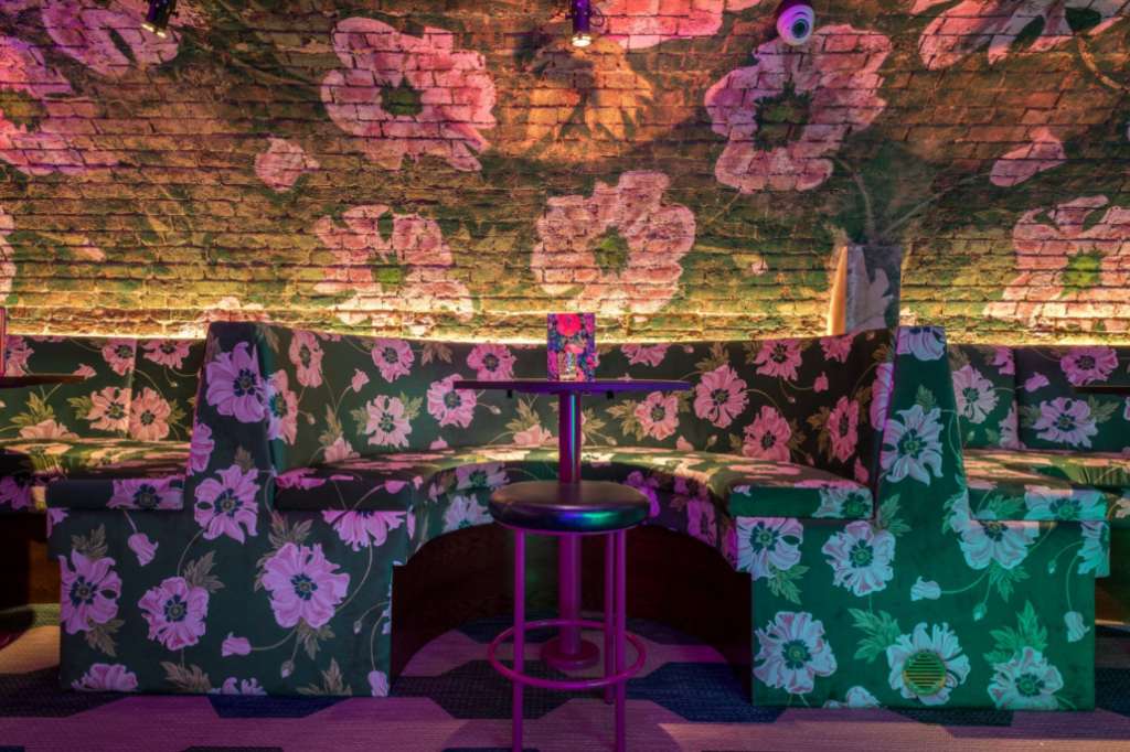 Back bar with floral-patterned booth seating, high-top table, and pink-purple ambient lighting on brick walls.