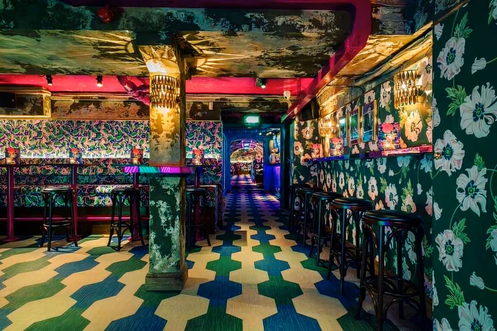 Back bar with floral wallpaper, geometric floor, bar seating, gold ceiling details, and colorful accent lighting.