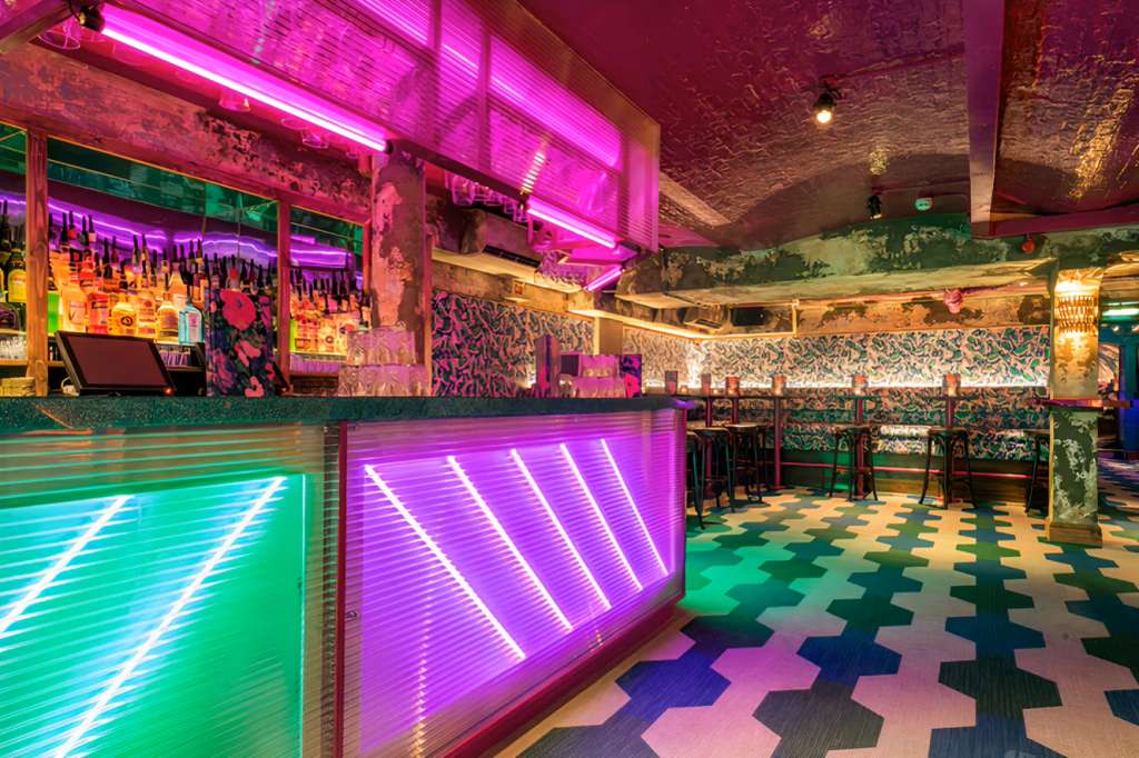Back bar with neon-lit counter, magenta and green lighting, patterned floor, and bottle-lined shelves.