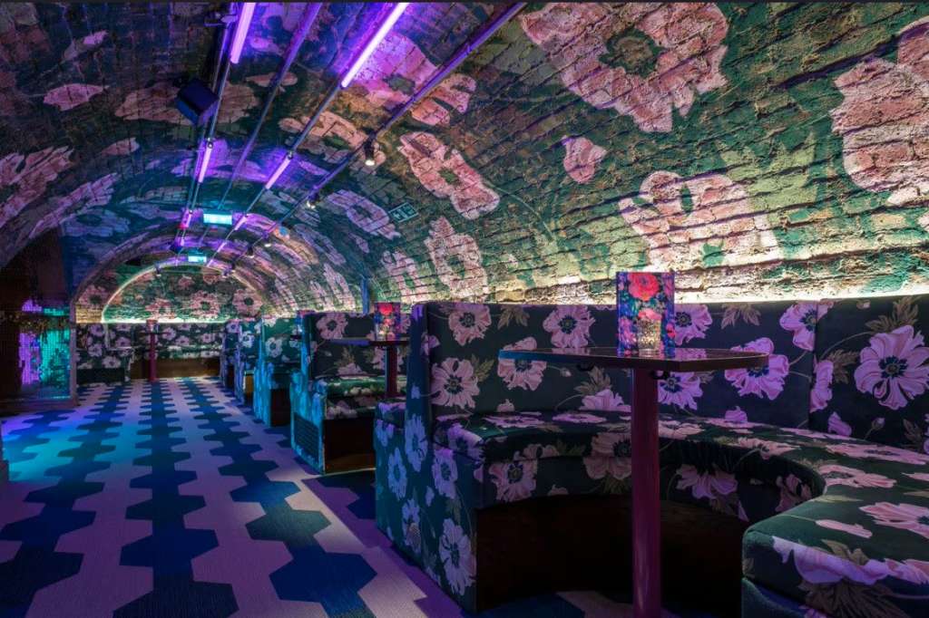 Arched brick corridor with purple neon lighting, floral wall projections, geometric floor pattern, and booth seating.