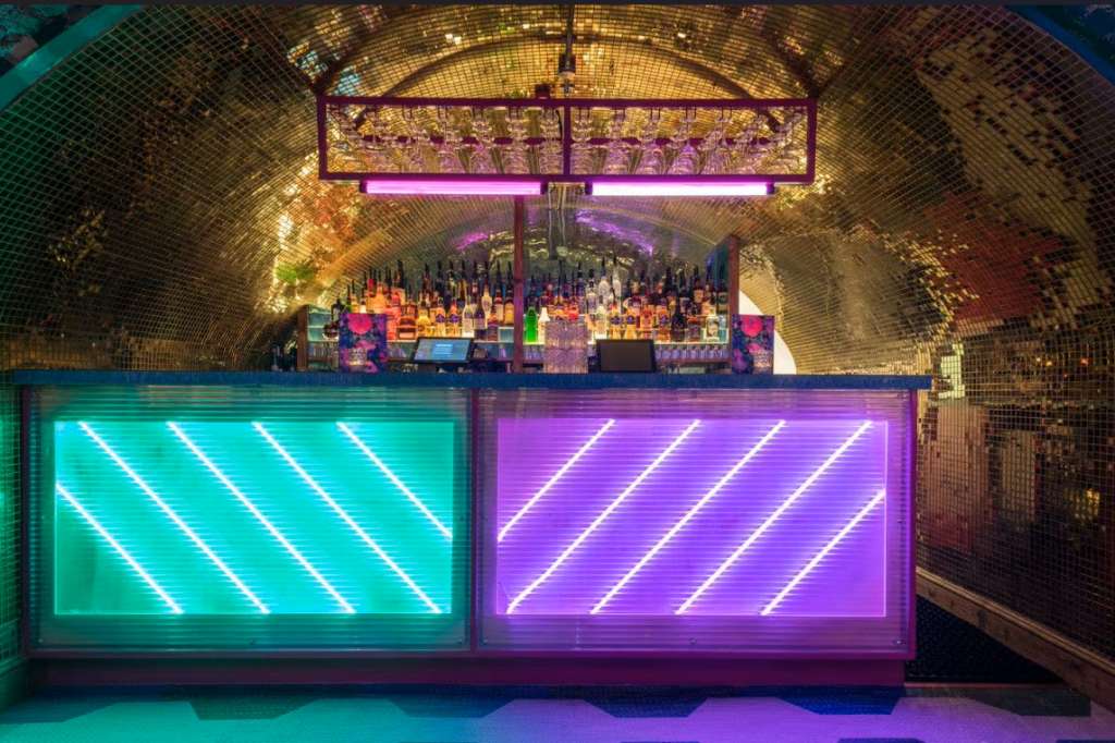 Back bar with neon-lit counter, tiered bottle display, and gold mosaic arched ceiling under colored lighting.