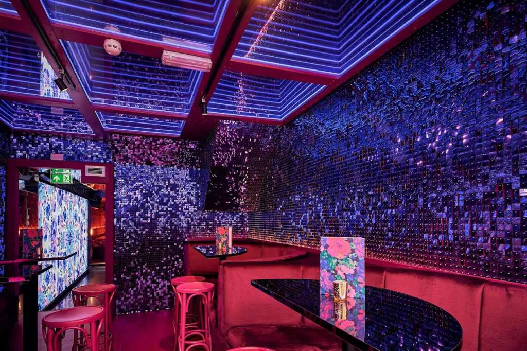 Modern bar lounge with neon blue and pink lighting, sequined walls, red seating, and digital screens.