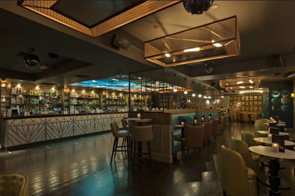 Modern bar and lounge with geometric bar front, booth seating, ambient lighting, and full-height spirits display.