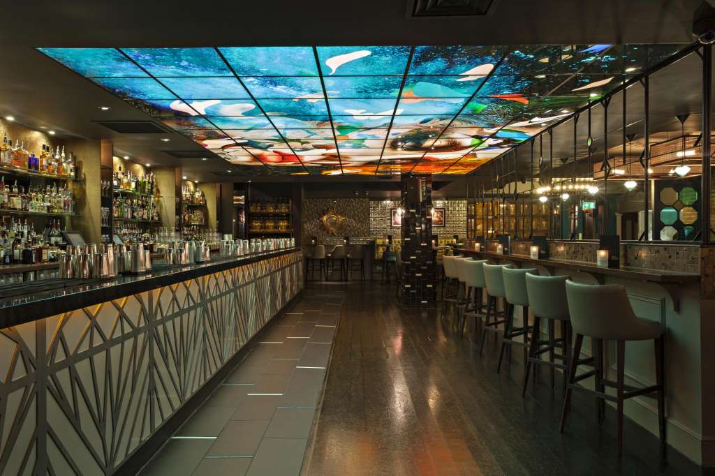 Modern bar interior with illuminated sky ceiling, extensive bottle display, geometric paneling, and mint-green bar seating.