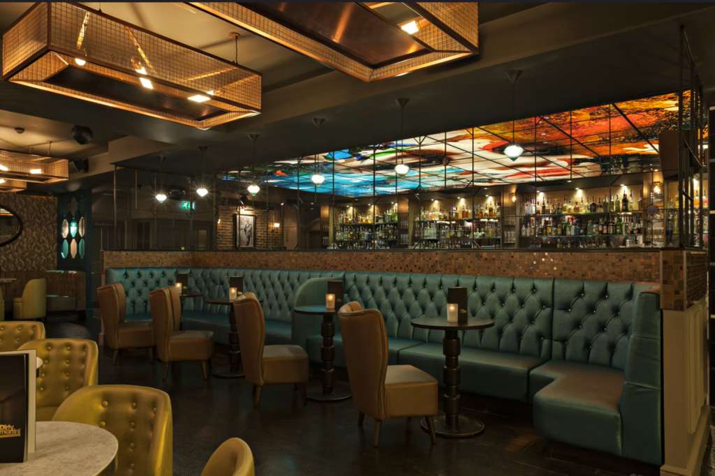 Upscale bar lounge with tufted green seating, brass fixtures, backlit artwork, and full-service bar counter.