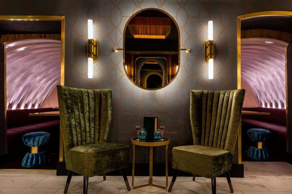 Luxury lounge with olive velvet chairs, gold-framed mirrors, geometric wallpaper, and ambient lighting.