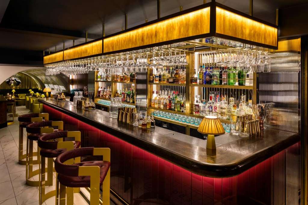 Modern bar with curved counter, burgundy and gold seating, extensive spirit bottles, and dramatic overhead lighting.