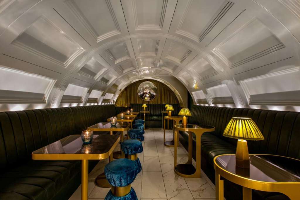 Art deco lounge with barrel ceiling, black banquettes, gold tables, brass lamps, and blue velvet chairs.