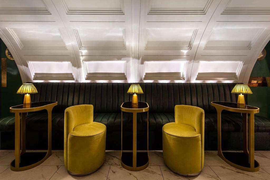Lounge area with black banquette seating, yellow velvet chairs, brass side tables, and brass lamps under coffered ceiling.