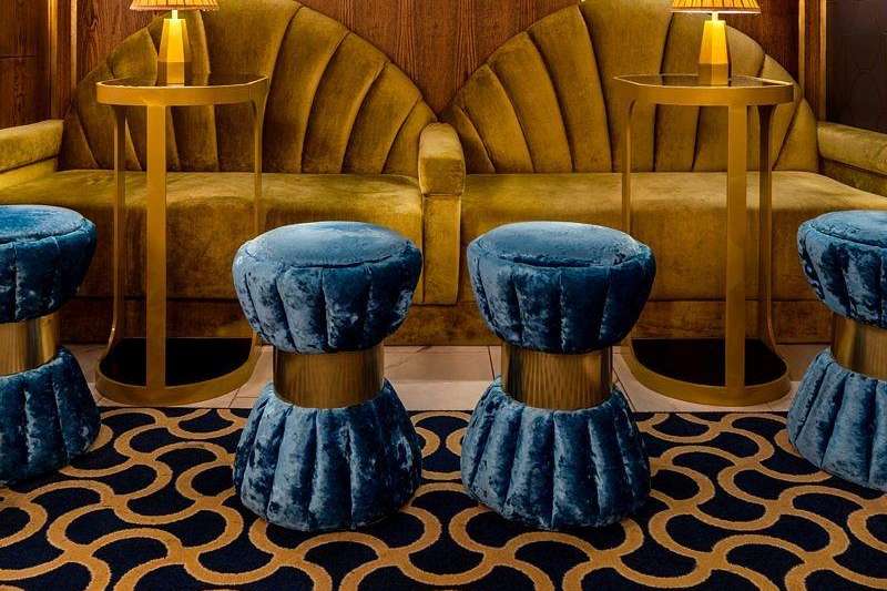 Upscale lounge with gold velvet seating, blue velvet stools, illuminated bar displays, and geometric patterned flooring.