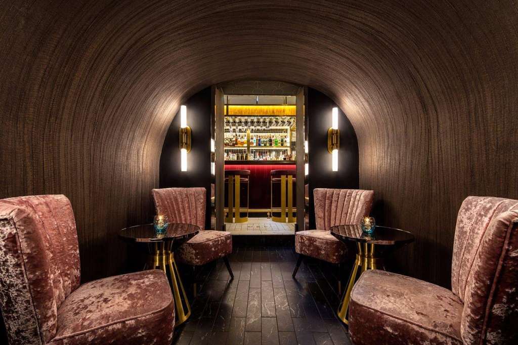 Luxurious bar lounge with curved brick walls, velvet seating, gold accents, and illuminated bar counter in background.