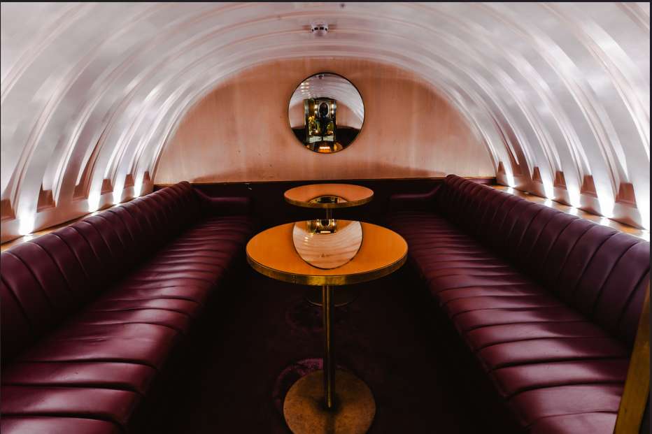 Narrow barrel-vaulted room with burgundy leather banquette seating, brass tables, and recessed lighting.