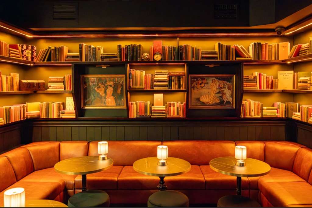 Intimate lounge with curved orange velvet seating, brass tables, candlelit ambiance, and illuminated bookshelf wall.