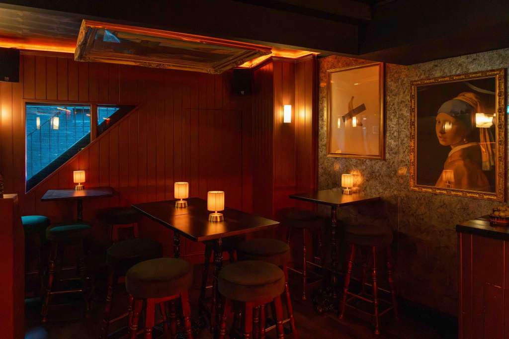 Intimate bar lounge with red wood paneling, high-top tables, bar stools, warm lighting, and framed artwork on walls.