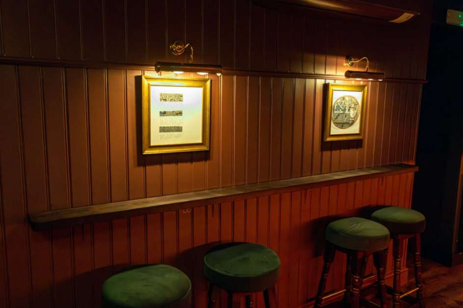 Bar counter with wood paneling, green velvet bar stools, and framed artwork on illuminated walls.