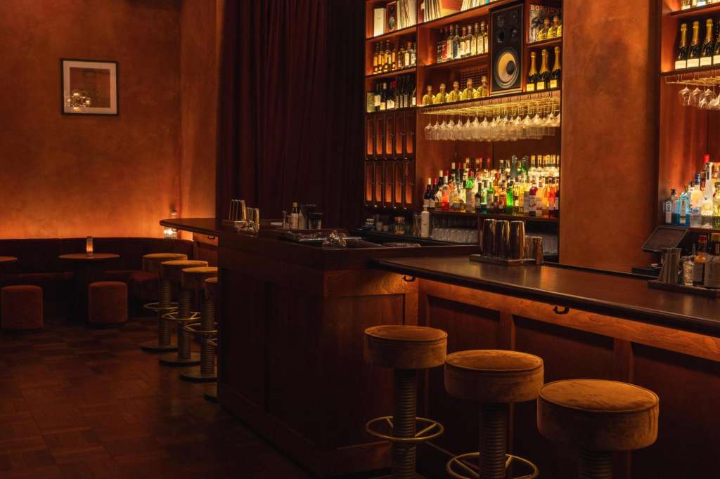 Bar interior with amber lighting, stocked shelves, bar seating, and decorative pendant lights.
