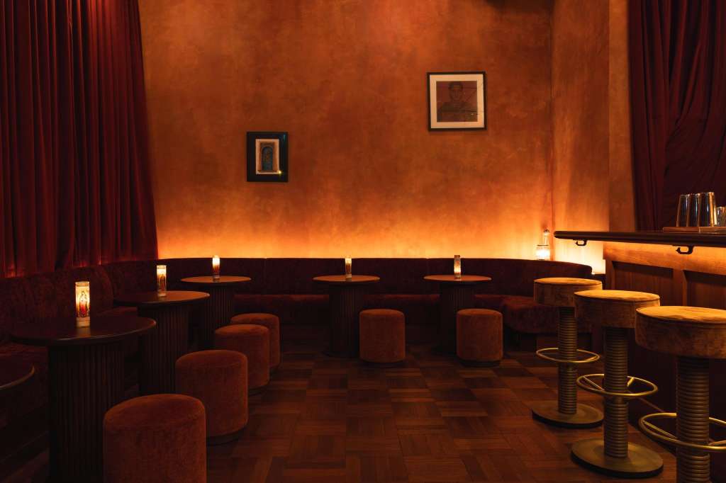 Upscale bar interior with warm amber lighting, red velvet curtains, dark seating, and framed artwork on terracotta walls.