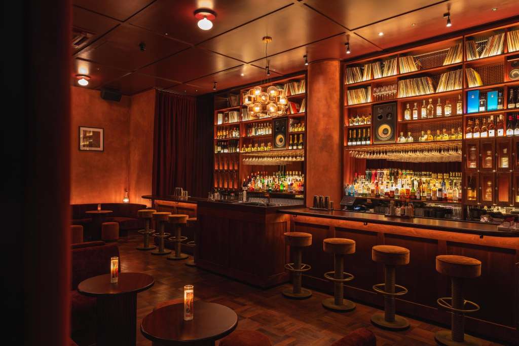 Bar interior with warm orange lighting, extensive bottle display, bar seating, and round tables with candles.