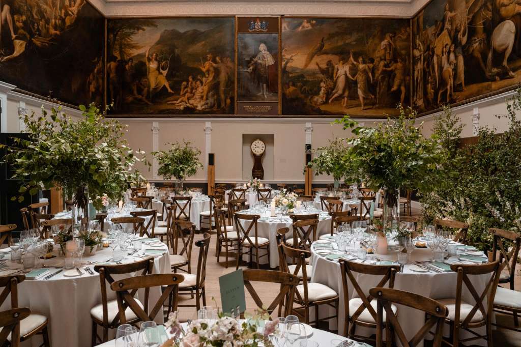 Great Room set for a formal dinner with round tables, white linens, wooden chairs, and tall floral arrangements beneath classical paintings.