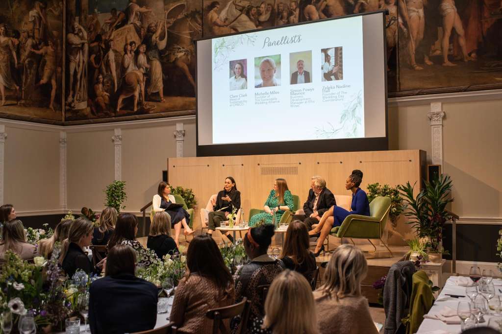 Great Room set up for a panel discussion with five speakers seated on stage, projection screen displaying panelists, and audience seated at tables with floral arrangements.