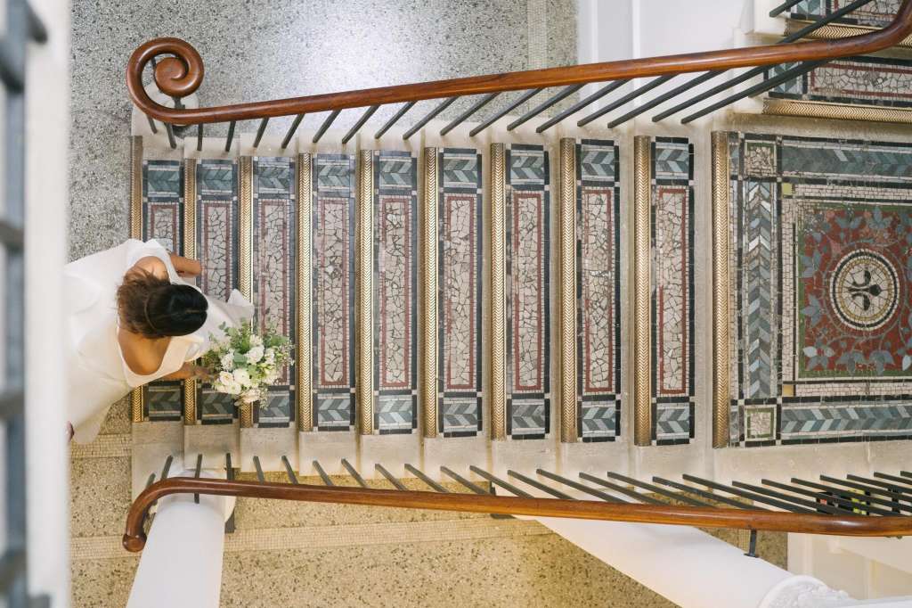 Bride in white dress standing on ornate mosaic floor with decorative staircase railing and classical architectural details.