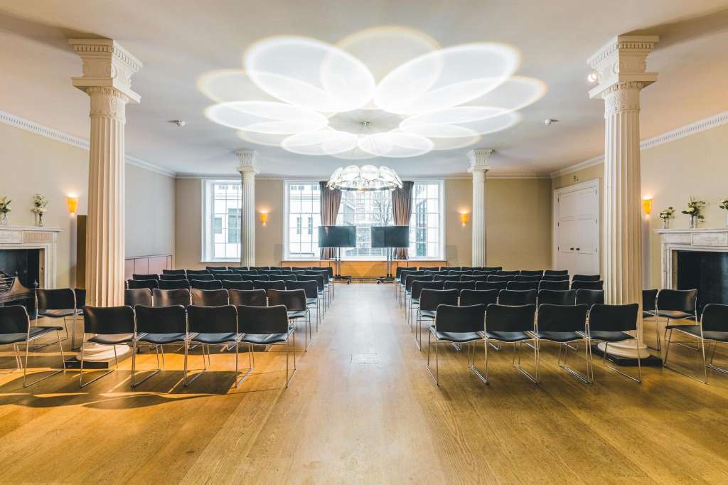 Great Room set up with rows of black chairs facing a stage, ornate columns, and modern ceiling lighting.
