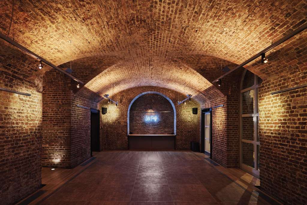 Vaulted brick corridor with arched ceilings, warm uplighting, and dark flooring. Empty event space.