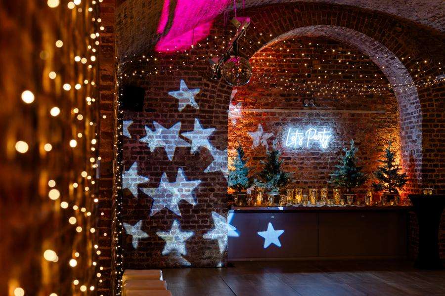 Vaulted brick room decorated with blue star projections, warm string lights, neon signage, and small evergreen plants.