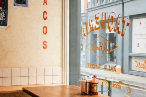 Casual dining space with wooden table, yellow chair, and street-facing window with signage.