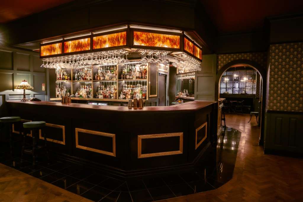 Bar lounge with curved counter, illuminated amber-glass ceiling feature, and extensive bottle display behind backlit shelving.