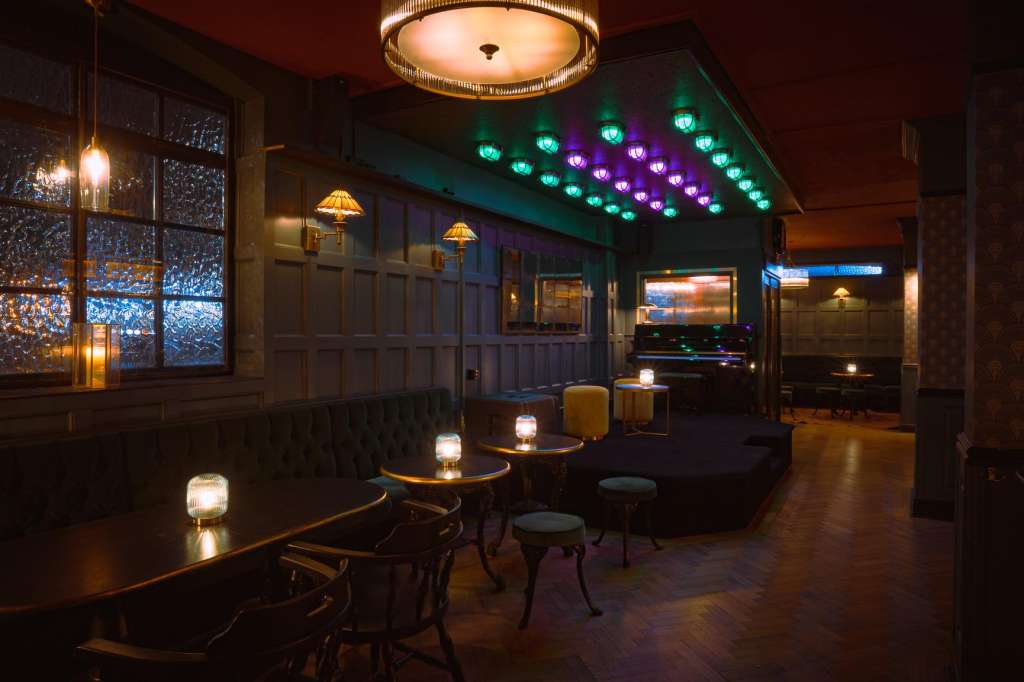Lounge interior with round tables, ambient lighting, green and purple accent lights, and large windows overlooking city lights.