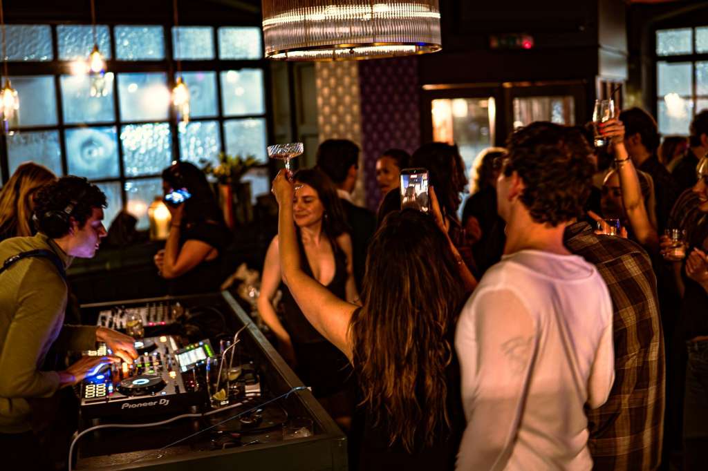 Evening reception with DJ booth, guests mingling and holding drinks under pendant lighting and large windows.