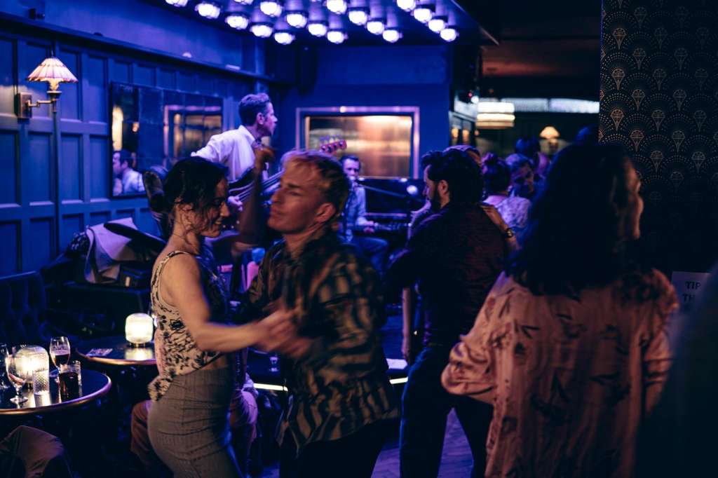 Bar lounge with guests mingling under blue ambient lighting, round tables with drinks and candles.