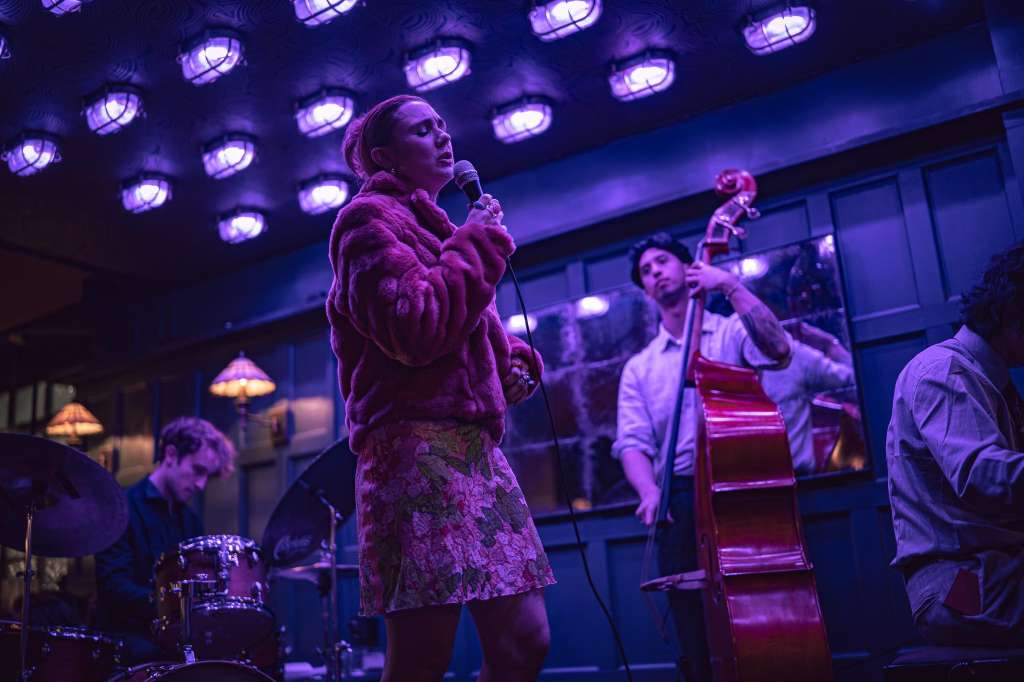 Live band performance with singer at microphone, drummer, and cellist under purple stage lighting.