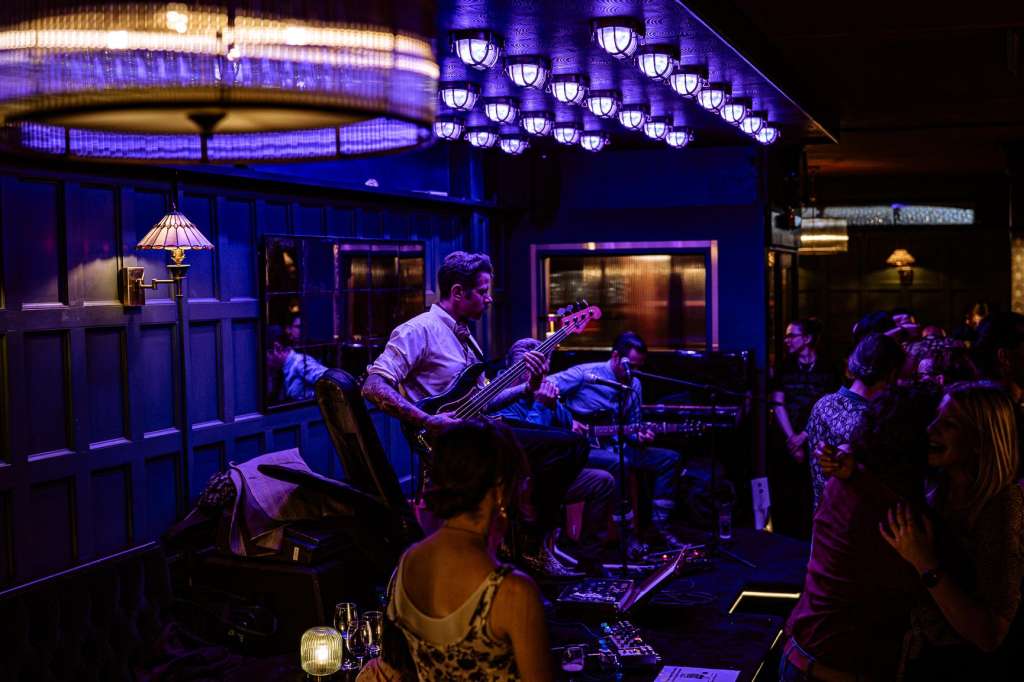 Bar lounge with live band performing, purple ambient lighting, guests seated and standing around performance area.