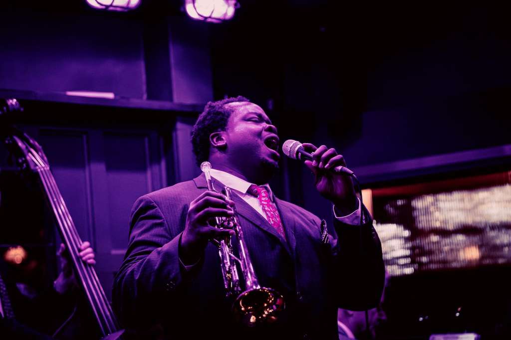 Live jazz performance with musicians playing saxophone and bass under purple stage lighting in an indoor venue.