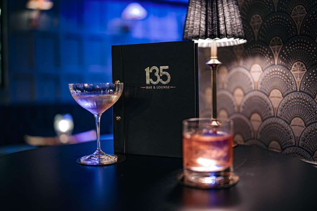 Bar counter with cocktails, branded signage, and decorative lamp against patterned backdrop.