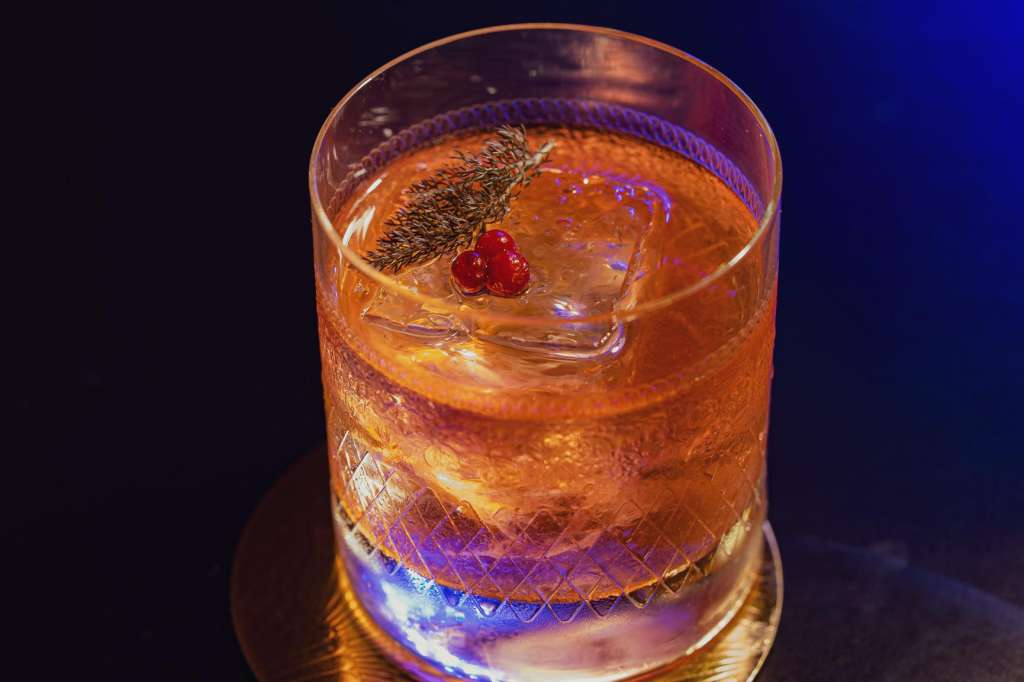 Craft cocktail with amber liquid, ice, red berries, and herb garnish on dark background.