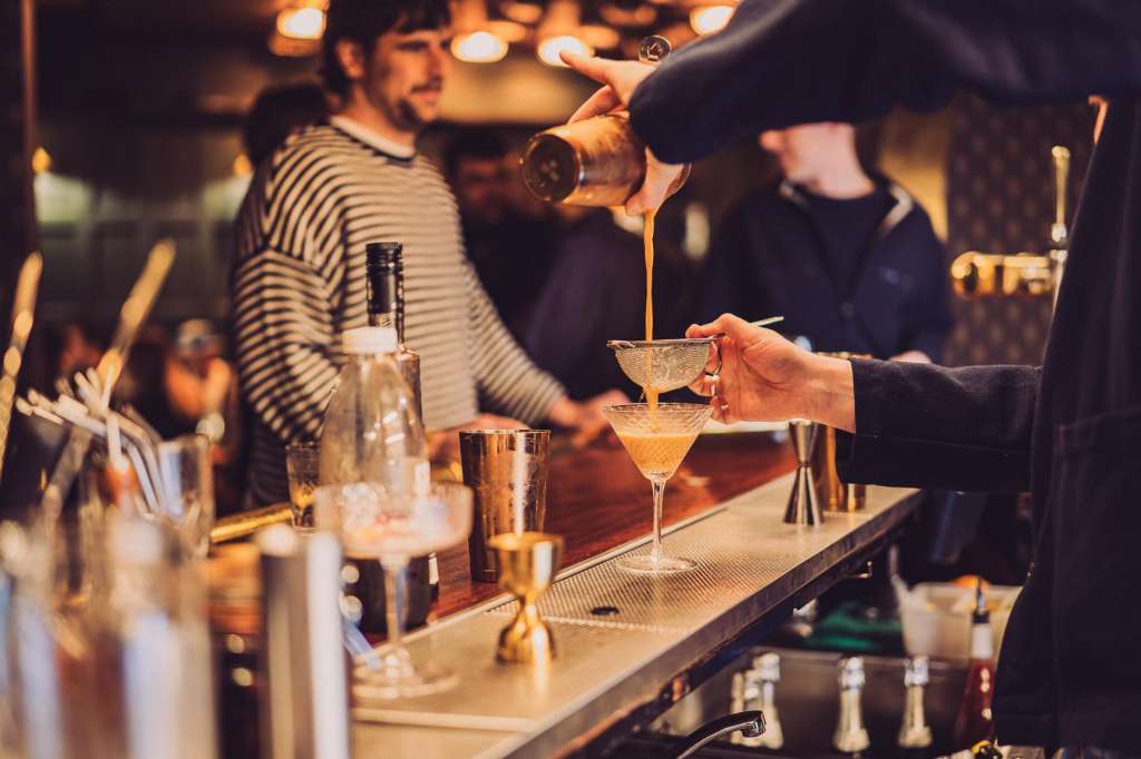 Bartender pouring cocktail at a bar counter with guests in warm ambient lighting.
