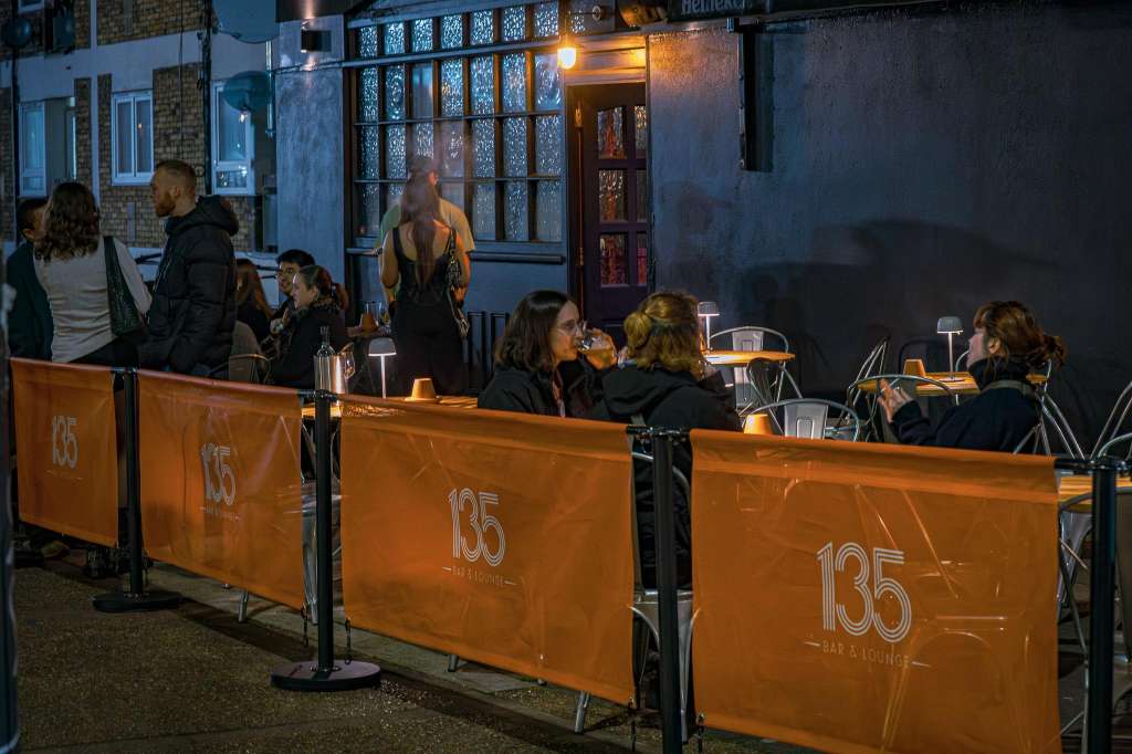 Outdoor terrace with branded barriers, guests seated at high tables with drinks and ambient lighting at dusk.