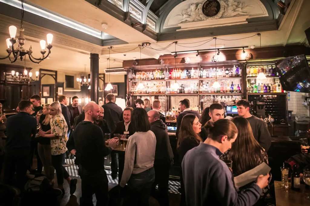 Bar interior with multiple guests standing and mingling, illuminated by chandeliers and bar lighting.