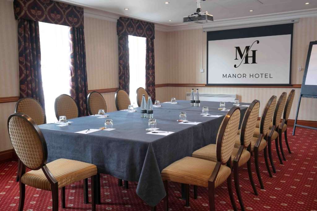 Meeting room set up with long rectangular table, blue tablecloth, place settings, and projection screen displaying Manor Hotel branding.
