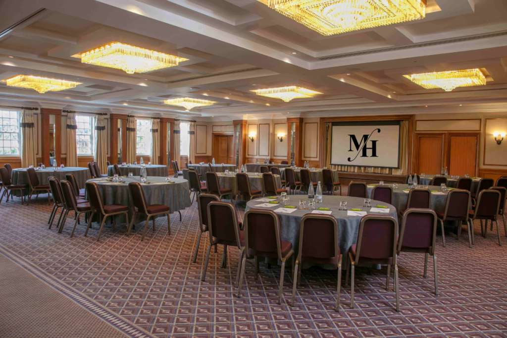 Ballroom set with round tables, burgundy chairs, grey linens, and crystal chandeliers overhead.