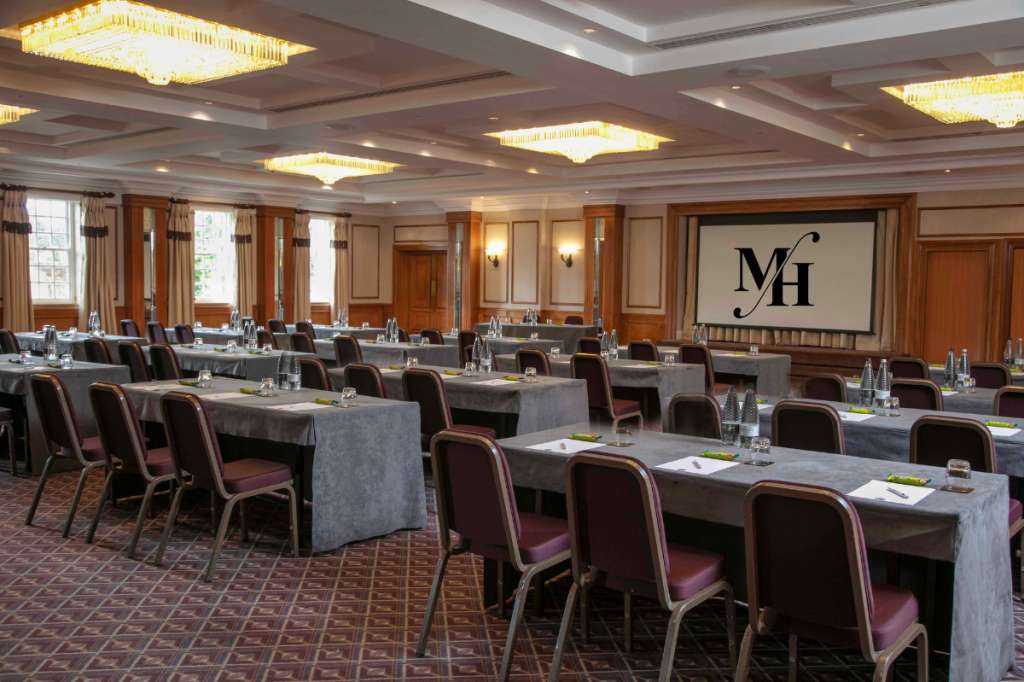 Conference room with long tables, burgundy chairs, projection screen, and decorative ceiling fixtures.