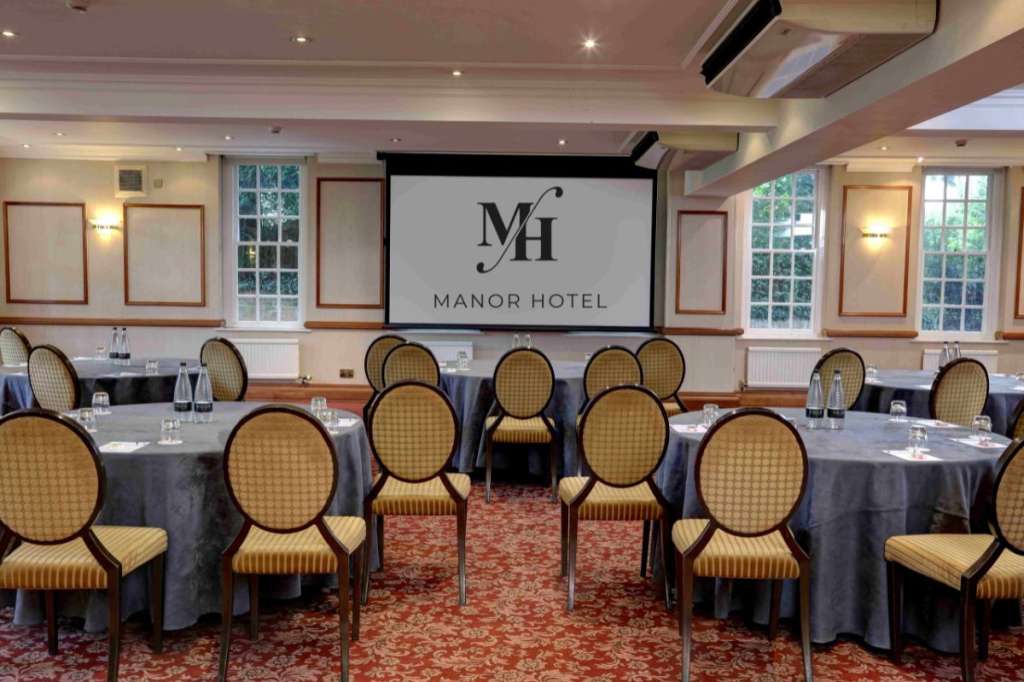 Banquet room with round tables dressed in navy linens, gold-patterned chairs, and projection screen displaying Manor Hotel logo.