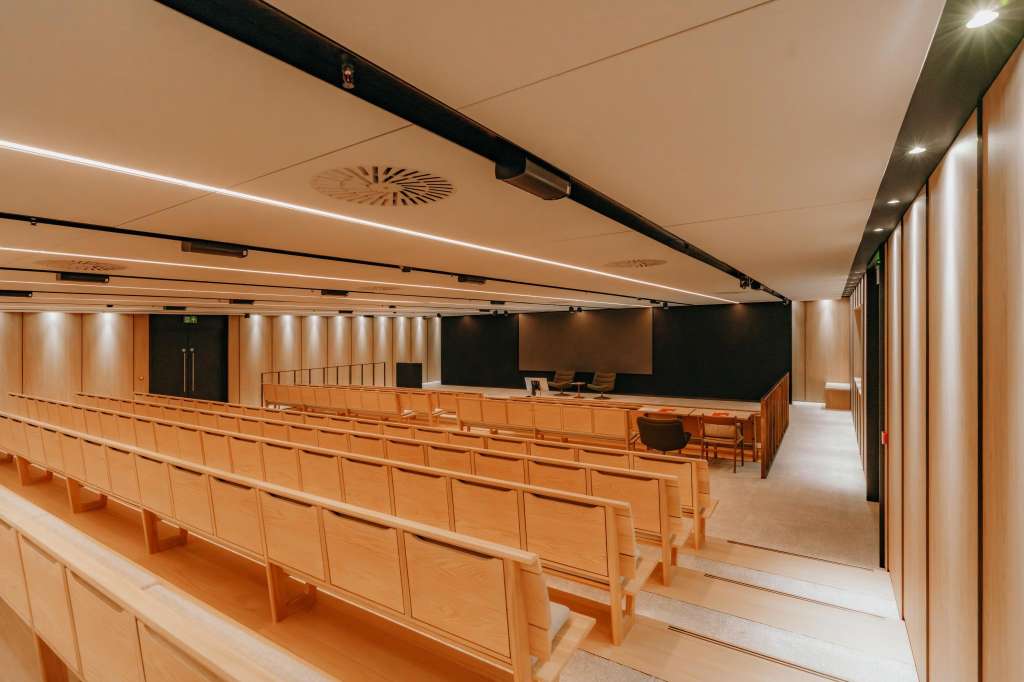 Auditorium with rows of wooden chairs facing a stage with black backdrop and presentation screens.