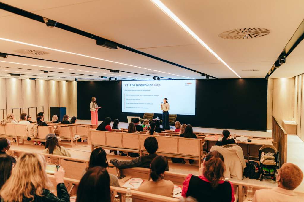 Atrium lecture hall with tiered seating, two speakers presenting at podium, large projection screen displaying presentation slide, audience seated throughout.