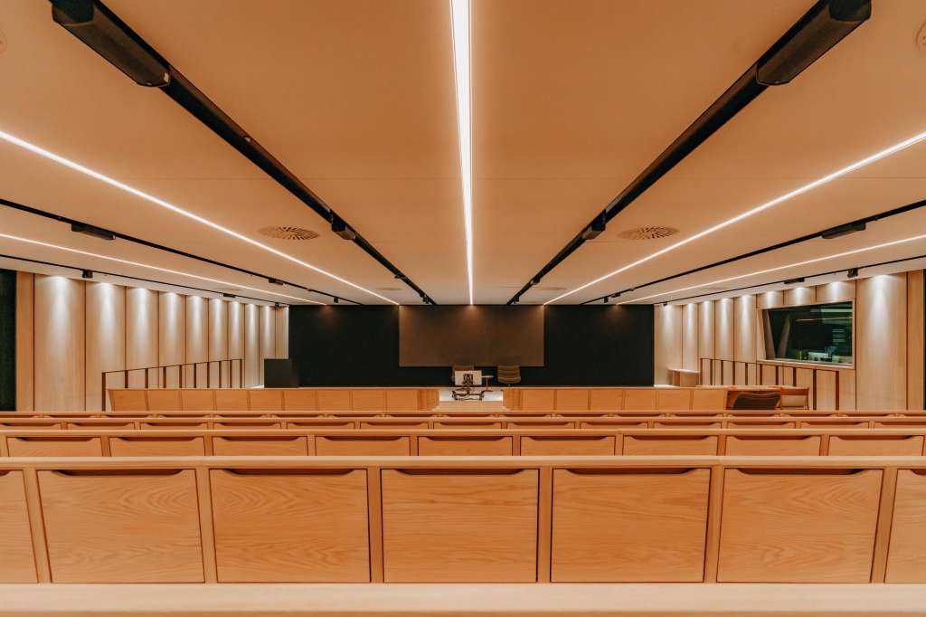 Atrium with tiered wooden seating, black acoustic panels, and linear ceiling lights facing a presentation stage.